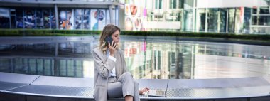 Şehir merkezinde dizüstü bilgisayarı ve cep telefonuyla oturan meşgul bir kadının portresi, kurumsal bir bayan birini arıyor ve bilgisayardaki işi kontrol ediyor..