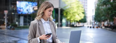 Şehir merkezinde çalışan takım elbiseli kadın dizüstü bilgisayar ve cep telefonu kullanıyor..