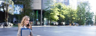 Ofis binasının yanında oturan genç bir kadının dikey görüntüsü, elinde laptop ve iş belgeleriyle birlikte klasörler, cep telefonundan birini ararken..