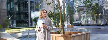 Takım elbiseli güzel bir iş kadını, sokakta hazır kahve içiyor, yemek molasında ofis binasının yakınında birini bekliyor..