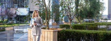 Takım elbiseli şık bir kadın, elinde belgelerle sokakta duruyor, iş bilgisayarı ve kahveyle iş merkezinin yakınında bekliyor..