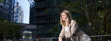 İş kadınının dikey görüntüsü, iş merkezinin önünde duruyor, cep telefonuyla konuşuyor..