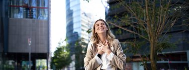 Hevesli iş kadını seviniyor, kutlama yapıyor ve mutlu hissediyor, bej takım elbisesiyle sokakta dikilerek saf neşeyi ifade ediyor..