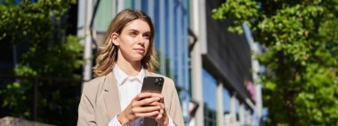 Şirket kıyafetleri içinde şık bir iş kadınının portresi, elinde akıllı telefon, sokakta duruyor ve elinde cep telefonu kullanıyor..