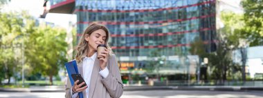 İşkadını sokakta kahve içerken, bej takım elbiseyle, elinde belgeler ve cep telefonuyla dikey çekim yapıyor..