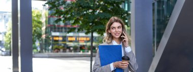 Meşgul iş kadını kahve içiyor, cep telefonuyla konuşuyor, iş belgelerini tutuyor, şehir merkezinde ofise yakın yürüyor..