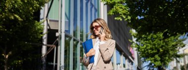 Genç başarılı iş kadını cep telefonuyla konuşuyor, iş belgeleriyle telefon ve dosya tutuyor, caddede ofisine yürüyor..