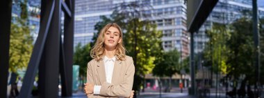 Genç satış elemanı portresi, takım elbiseli kendine güvenen bir iş kadını, göğsünde çapraz kollar, ofis binalarının yanındaki sokakta iktidar pozu..