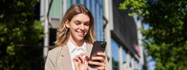 Gülümseyen güzel iş kadınının sokakta cep telefonunu kullanmasının portresi. Şirket kadını akıllı telefonunu dinliyor, mesajlaşıyor, sokakta dikiliyor..