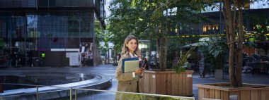 Kurumsal takım elbise giymiş, elinde laptop ve belgeler olan, kameraya gülümseyen güzel bir kadın..