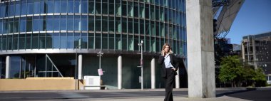 Takım elbiseli genç bir kadın sokakta yürüyor ve cep telefonuyla konuşuyor. İş kadını işe giderken telefonla konuşuyor..