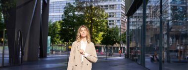 Kendine güvenen bej takım elbiseli genç bir iş kadını portresi, şirket avukatı ya da satış görevlisi caddede ofis binasının önünde duruyor ve gülümsüyor..