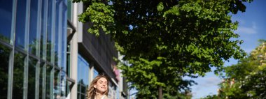 Dosyası olan bir iş kadınının dikey resmi ofis binalarının yakınındaki bir sokakta duruyor, ofis işi için takım elbise giyiyor..