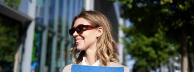 Güneş gözlüklü, takım elbiseli, iş belgeleriyle dolu bir klasörü olan, iş görüşmesine giden, dışarıda duran gülümseyen genç bir kadının yakın portresi..