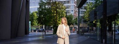 Kendine güveni tam, bej takım elbiseli genç bir kadının portresi, iş kıyafetleriyle caddede duruyor, kendinden emin görünüyor..