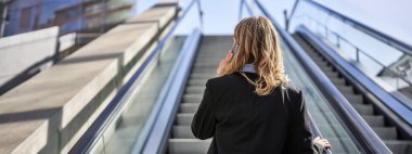 Takım elbiseli bir iş kadınının yürüyen merdivende dikilip cep telefonuyla konuşması..