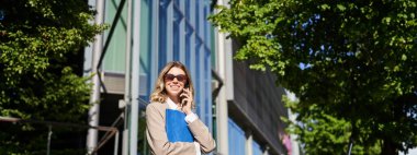 Güneş gözlüklü çalışan bayan, ofis binasının önünde cep telefonuyla konuşan satıcı, sokakta duran, klasörü tutan..
