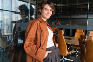 Kendine güvenen genç iş kadını portresi, ofisinde poz veren dijital göçebe, günlük kıyafetler giymiş ve gülümsüyor..