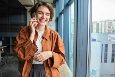 Genç kadın çalışanın görüntüsü, yönetici cep telefonuyla konuşuyor, pencerenin yanında duruyor ve gülüyor, birini arıyor, telefonla konuşuyor..