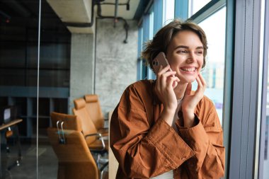 Genç kadın çalışanın görüntüsü, yönetici cep telefonuyla konuşuyor, pencerenin yanında duruyor ve gülüyor, birini arıyor, telefonla konuşuyor..