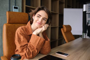 Genç bayan girişimcinin yakın portresi, günlük giysiler içinde satış elemanı, ofisinde oturuyor ve gülümsüyor..