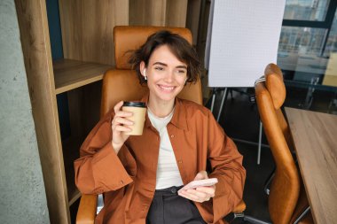 Ofisinde çalışan, elinde akıllı telefon tutan, kahve içen ve gülümseyen, öğle yemeği molasından keyif alan kadın girişimcinin portresi..