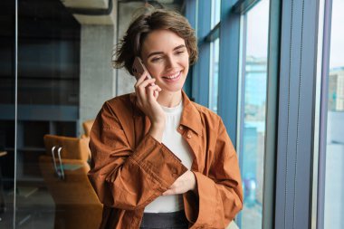 Pencerenin yanında genç ve mutlu bir ofis çalışanı, birini arıyor, cep telefonuna cevap veriyor, konuşuyor ve gülümsüyor..