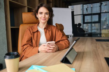Kendine güveni tam genç bir kadının portresi, ofiste çalışan günlük giysiler içindeki işverenin tabletinde dijital tablet ve kahvesi var, kendine güvenen yüz ifadesiyle gülümsüyor..