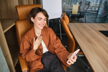 Günlük giysiler içinde bir kadın, yönetici ofisinde oturuyor ve çalışıyor, online toplantısı var, akıllı telefon uygulamasından görüntülü sohbete bağlanıyor..