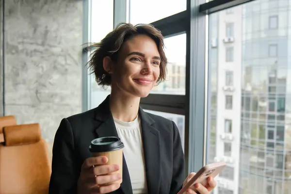 Portrait of corporate woman reading message on mobile phone, drinking coffee, standing near office mirror and smiling.
