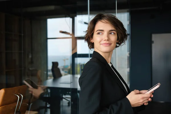 Portrait of corporate woman in suit, standing in office room with smartphone and smiling.