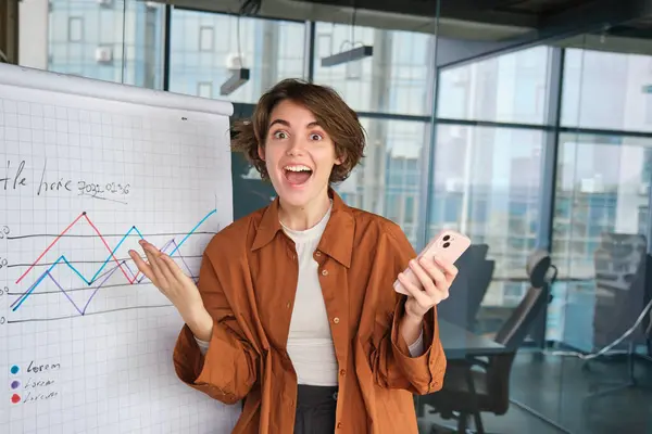 Portrait of excited ceo manager, business woman reacts amazed to diagram with company statistics, holds smartphone, celebrates victory.