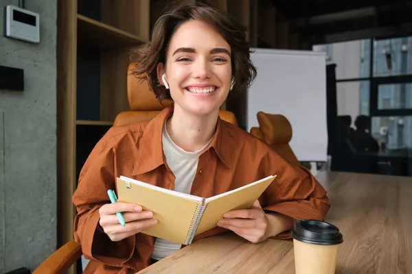 Portrait of young cheerful woman working on start up project, making notes, holding pen and planner, reading documents, sitting in an office.