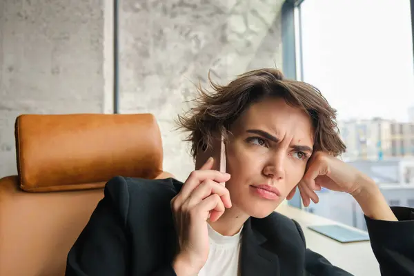 Close up portrait of working woman in an office, having difficult conversation, frowning while answering phone call, sitting in suit.
