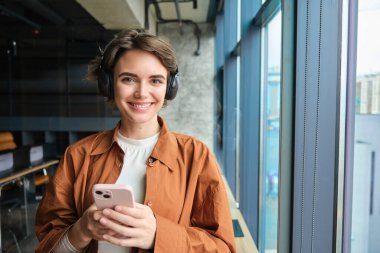Ofisteki genç modern kadın portresi, kablosuz kulaklıklı dijital göçebe, iş yerinde akıllı telefonuyla duruyor ve gülümsüyor, pencerenin yanında poz veriyor..