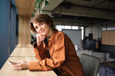 Genç, modern bir kadının resmi, ofiste çalışan, birlikte çalışan, çalışan, cep telefonu kullanan, kameraya mutlu bakan.