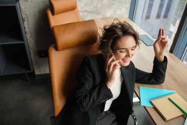 Satış elemanı telefonuna cevap veriyor, akıllı telefondan müşteriyle konuşuyor, bilgisayarlı bir ofiste çalışıyor, pencerenin yanında oturuyor..