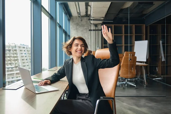 Portrait of young corporate woman, ceo manager in office saying hello to colleague, waving hand friendly, working on laptop near window, doing her job in coworking space.