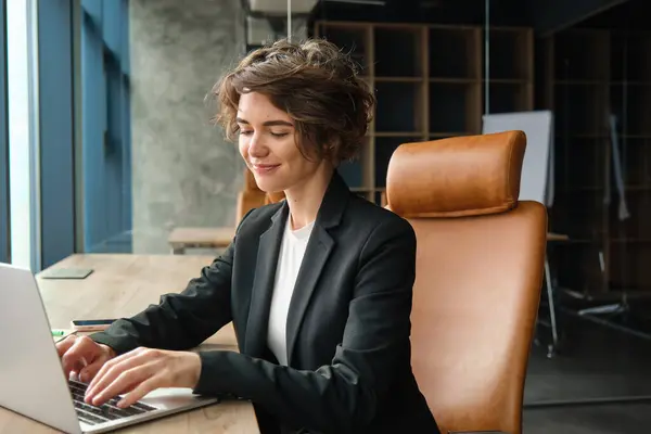 Portrait of woman working in her office. Businesswoman using laptop, messaging clients, replying to her coworker on team messenger, sitting in suit on chair near window.