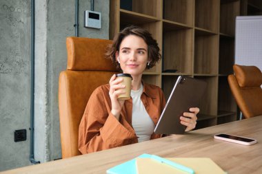 Ofiste genç bir kadının portresi, dijital tabletli girişimci, kahve içiyor ve gülümsüyor, proje üzerinde çalışıyor.