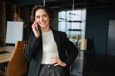 Kendine güveni tam bir satış elemanı portresi, cep telefonuyla konuşma, ofisinde cam duvarın ve konferans salonunun yanında dikilme, kameraya kendinden emin görünme..