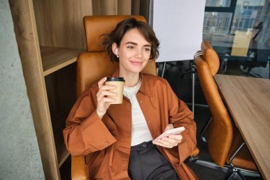 Ofisinde çalışan, elinde akıllı telefon tutan, kahve içen ve gülümseyen, öğle yemeği molasından keyif alan kadın girişimcinin portresi..