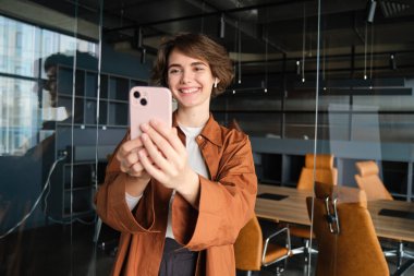 Gülümseyen güzel bir kadının portresi, ofis müdürü akıllı telefonuyla selfie çekiyor, şirket sosyal medyası için video kaydediyor, günlük kıyafetlerle poz veriyor..