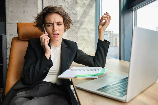 Portrait of frustrated saleswoman, businesswoman having an argument over the phone, difficult conversation over the telephone, sitting in office with laptop.