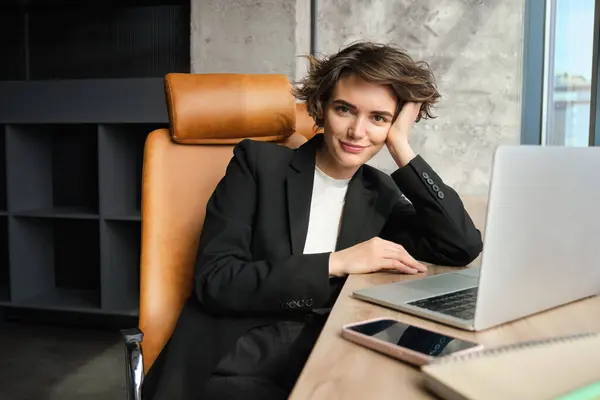 Young successful woman in business suit, sitting in her office corporation, lady boss working on laptop near window.