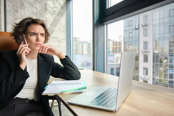 Portrait of serious woman having negotiation over the phone. Businesswoman with smartphone, looking at laptop screen with serious face, sitting in office.