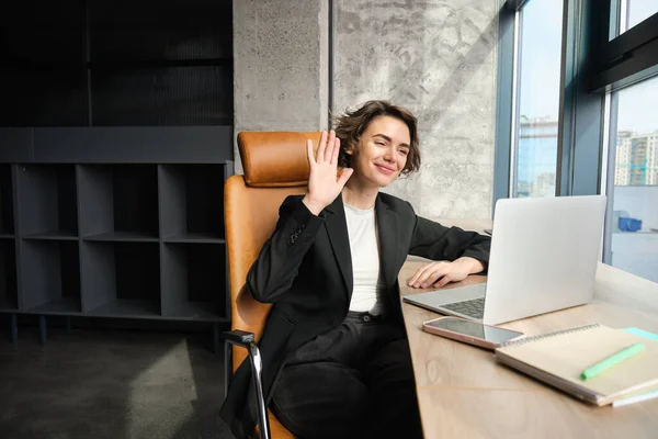 Portrait of corporate woman, ceo manager joins online meeting or webinar, saying hello at laptop camera, waving hand to greet coworkers, sitting in office.