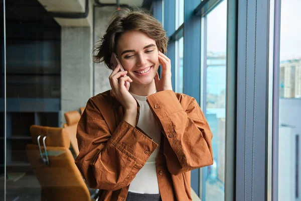 Portrait of working woman, social media manager in office, talking on mobile phone, having conversation on telephone, standing next to the window.