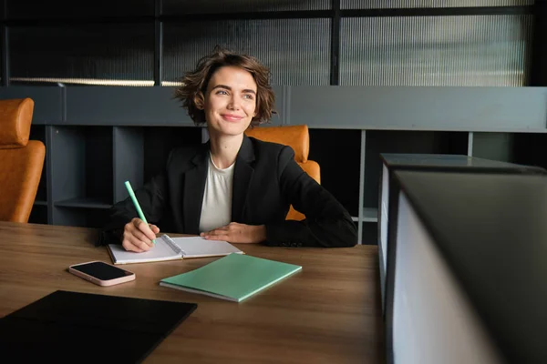 Portrait of successful saleswoman, corporate woman in suit, sitting in chair in her office with documents, writing down notes for meeting presentation, doing her job, looking outside window.