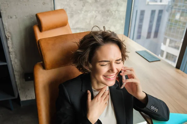 Portrait of business woman in suit, sitting in her office and answering a phone call with pleased smile, having a negotiation over the telephone conversation.